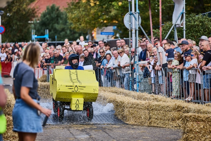 Een deelnemer aan de zeepkistenrace is zojuist van de schans afgerold en rijdt door de waterbak. Het publiek staat rijendik langs de kant. 